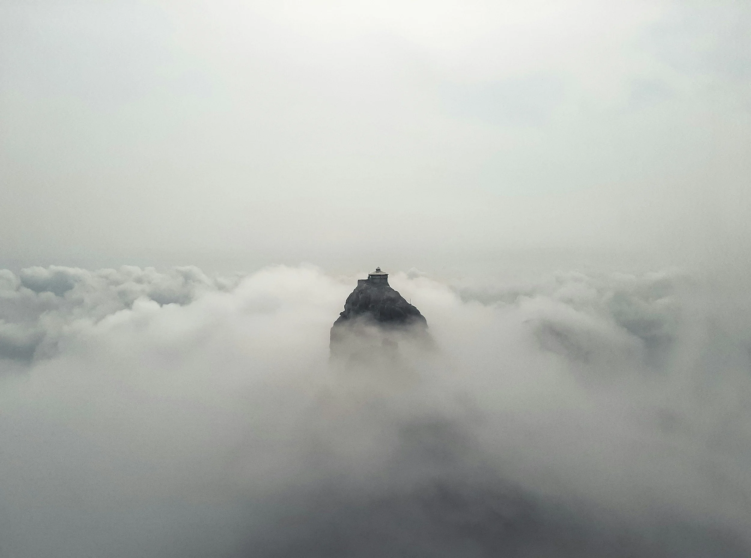 A mountain peak surrounded by clouds with a small building or temple on top, on a foggy day.