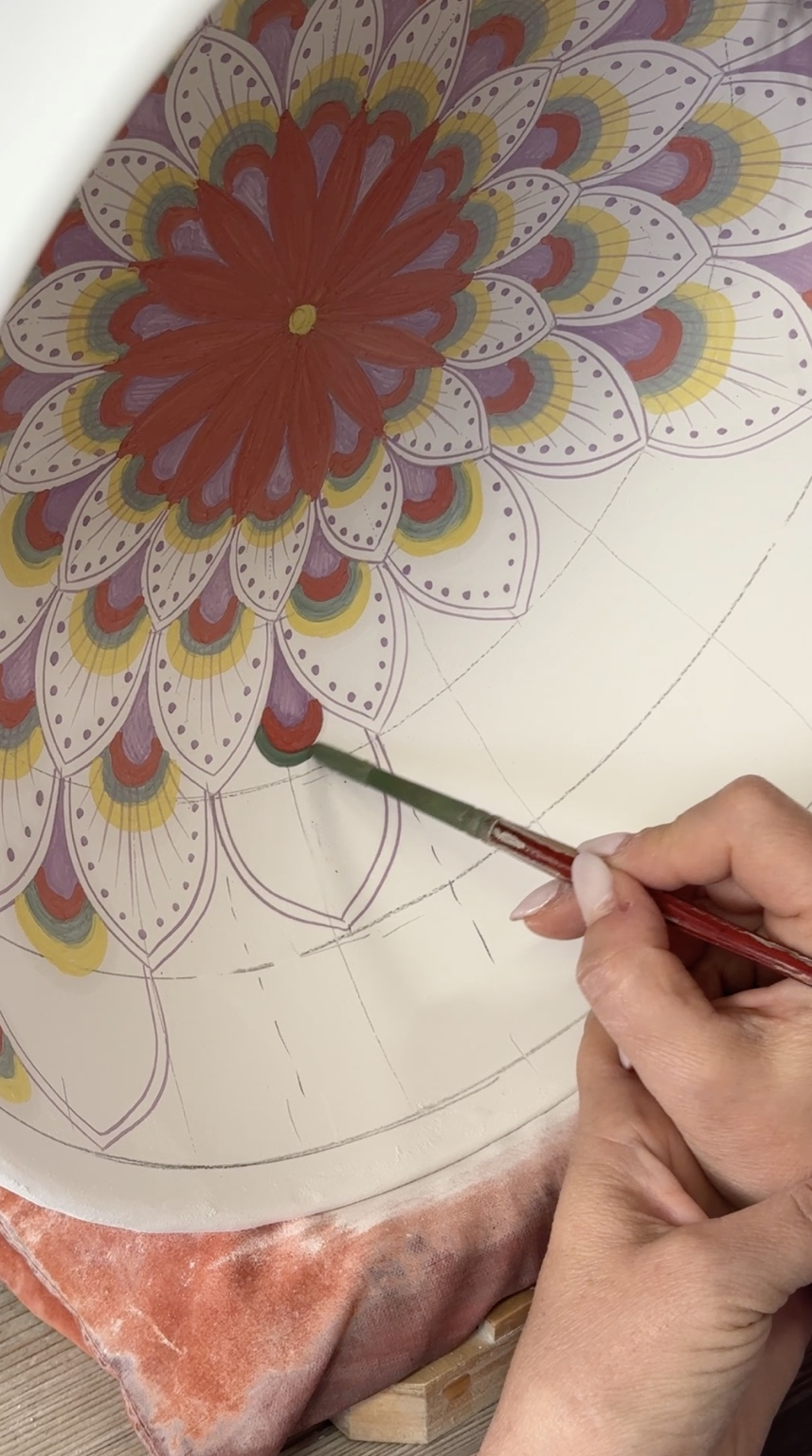 Artisan hand-painting Italian ceramic plate using brush with visible spacing guides and layered peacock pattern detail.