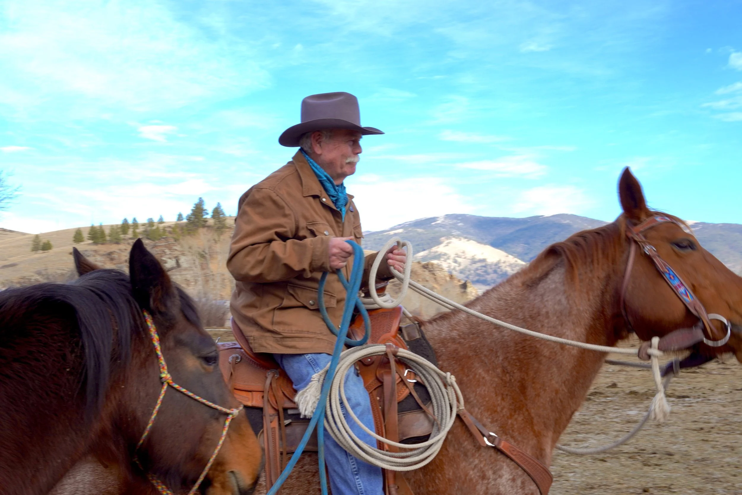 A man wearing a brown jacket and a cowboy hat rides a brown horse, holding the reins, with mountain hills and a partly cloudy sky in the background.