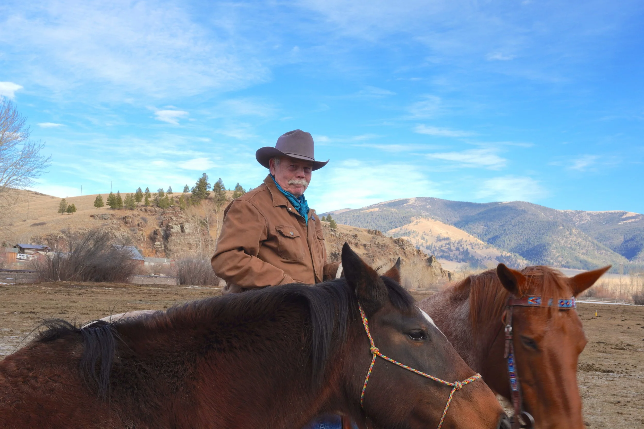 A man wearing a cowboy hat and brown jacket, standing with two horses in a rural landscape with mountains and a blue sky in the background.