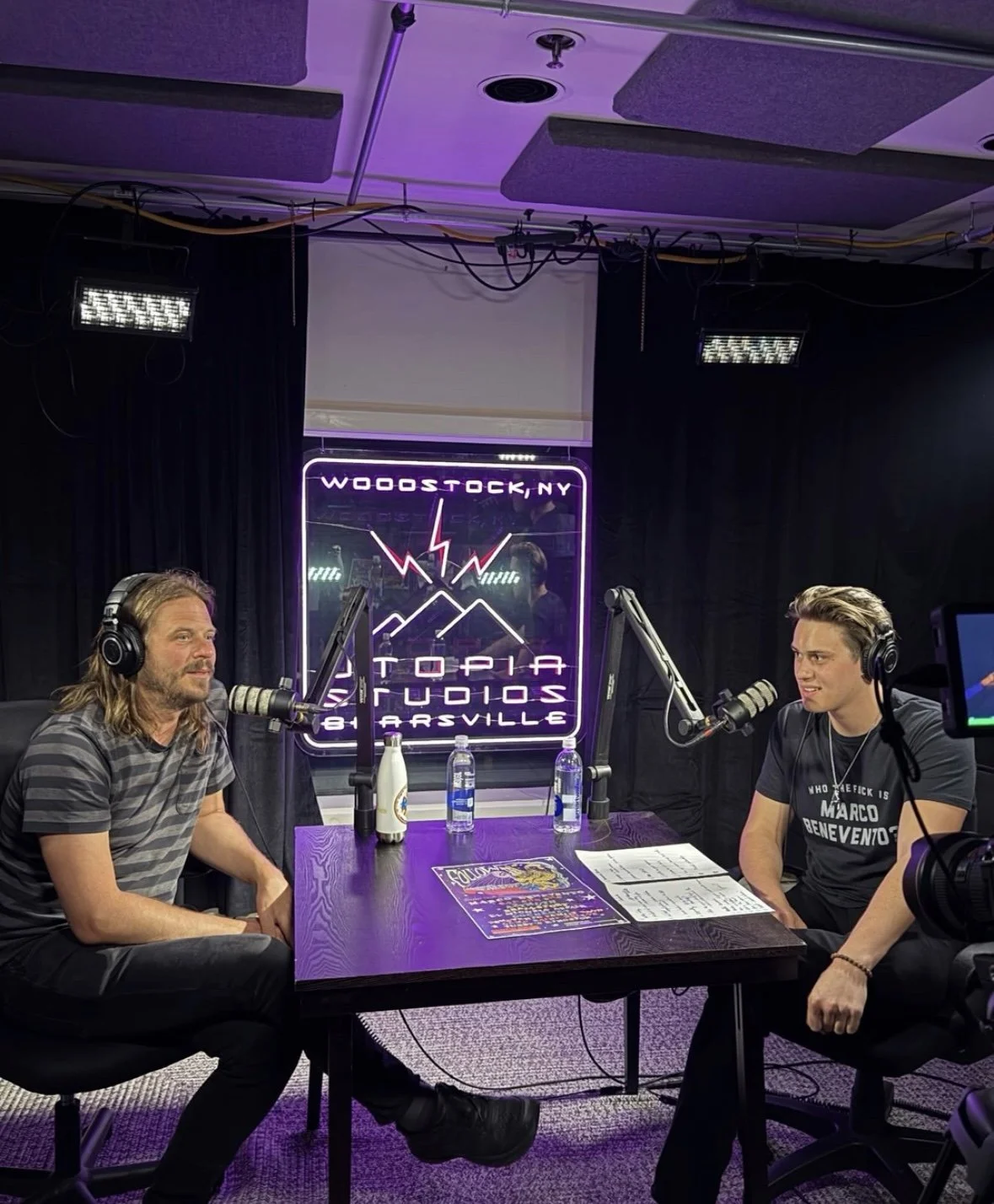 Two men are recording a podcast in a studio with black curtains, a table, and microphones. There is a neon sign in the background that reads "WooStock, NY, Utopia Studios, Bardville".