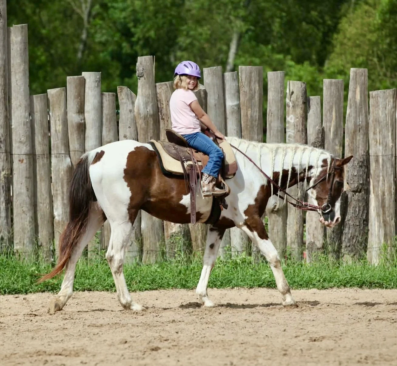 A young girl riding a brown and white pinto horse on a dirt path with a wooden fence and green trees in the background. She is wearing a purple helmet, pink shirt, and blue jeans.