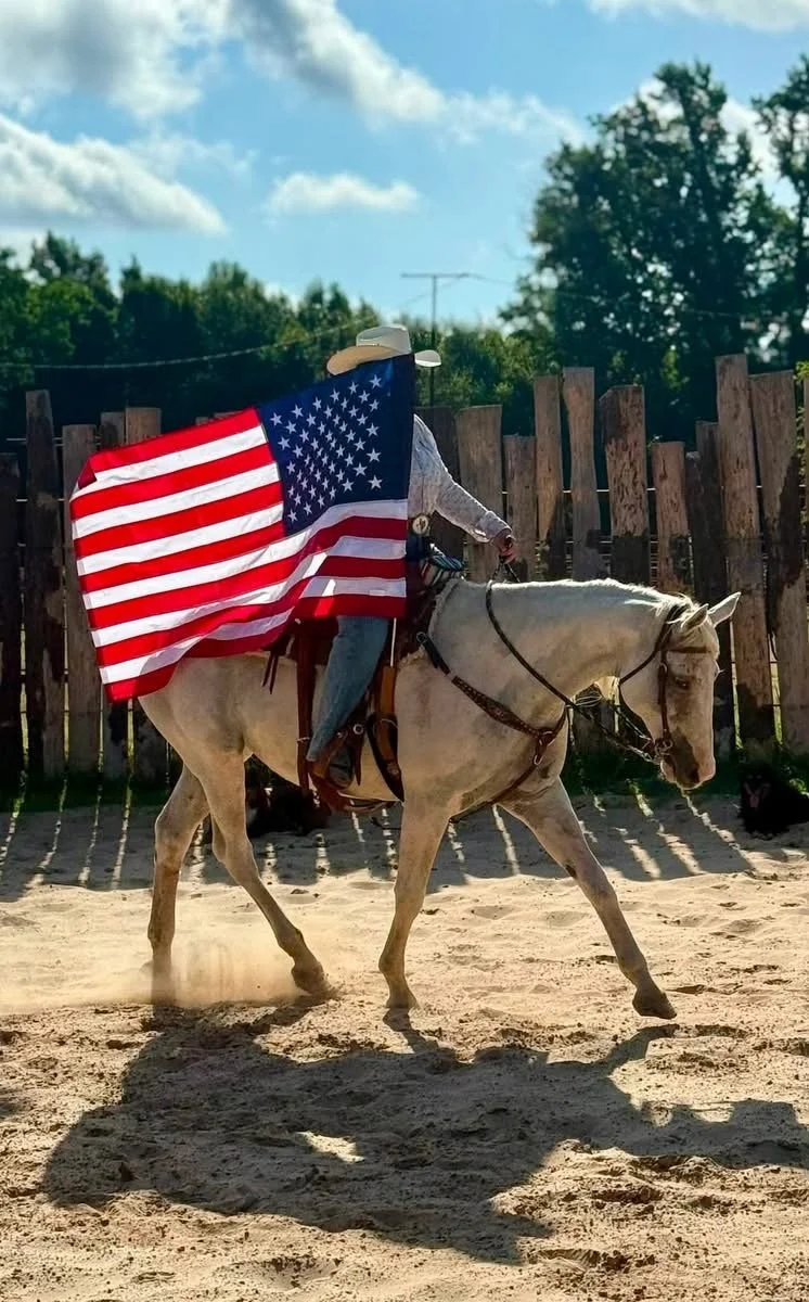 A person riding a white horse, carrying a large American flag, outdoors on a sunny day.