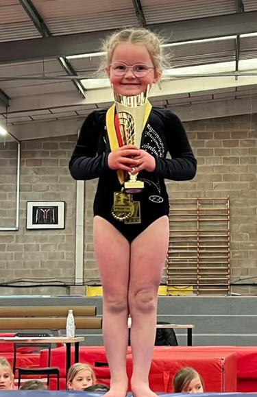 A young girl in a black gymnastics leotard standing on a red gymnastics mat, holding a large gold trophy, smiling with glasses, in an indoor gymnasium.