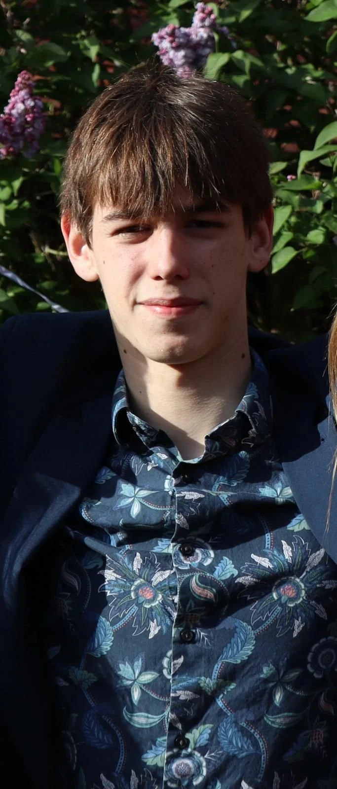 A young man with brown hair, wearing a blue floral patterned shirt and a dark jacket, stands outdoors in front of green foliage with purple flowers.