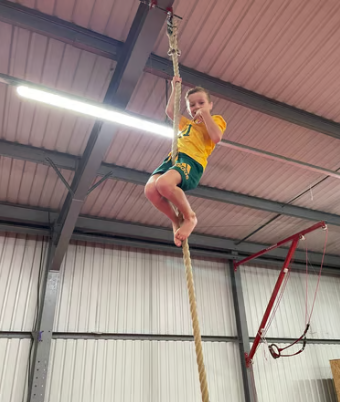 Young girl in yellow shirt and green shorts climbing a thick rope inside a gym, hanging from the ceiling