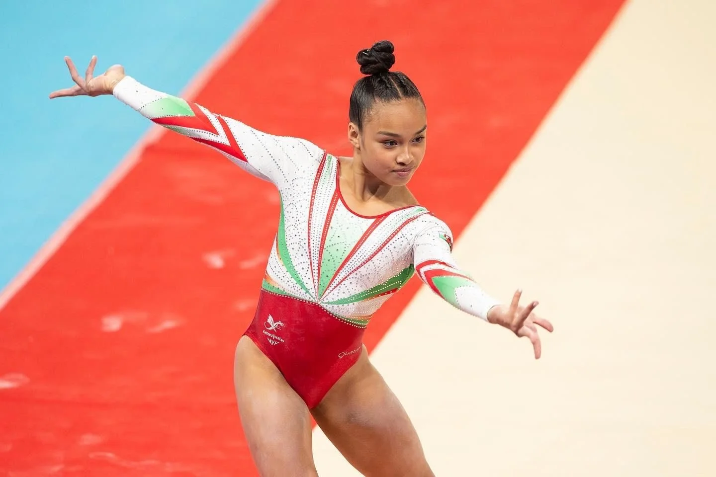 A female gymnast performing a routine on the balance beam, wearing a red, white, and green leotard with her hair styled in a high bun.