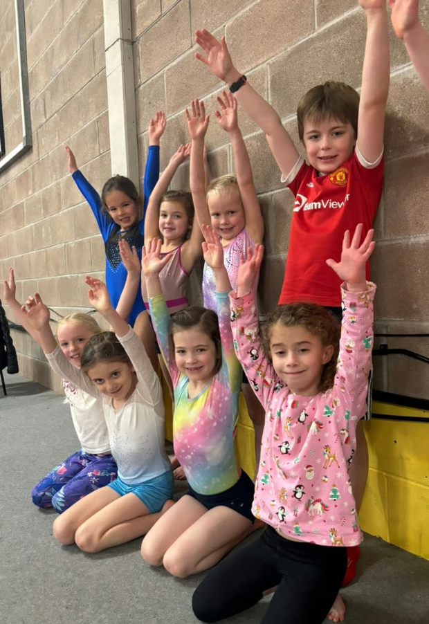 Seven children, six girls and one boy, pose with arms raised while sitting and standing against a brick wall in a gymnastics or dance studio.