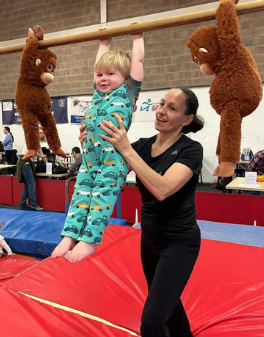 A woman assisting a young child in a gymnastics facility, holding the child up while hanging from a horizontal bar. Two people in bear costumes stand on the mat nearby.
