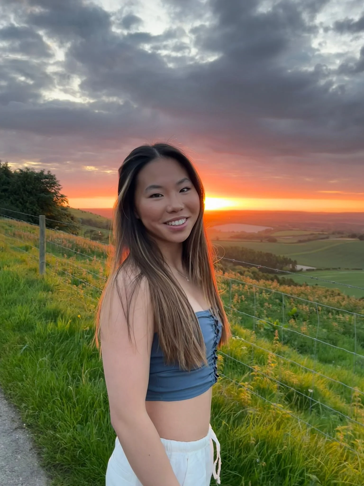 A woman with long brown hair smiling at sunset in a rural landscape with green fields, hills, a lake, and a cloudy sky.