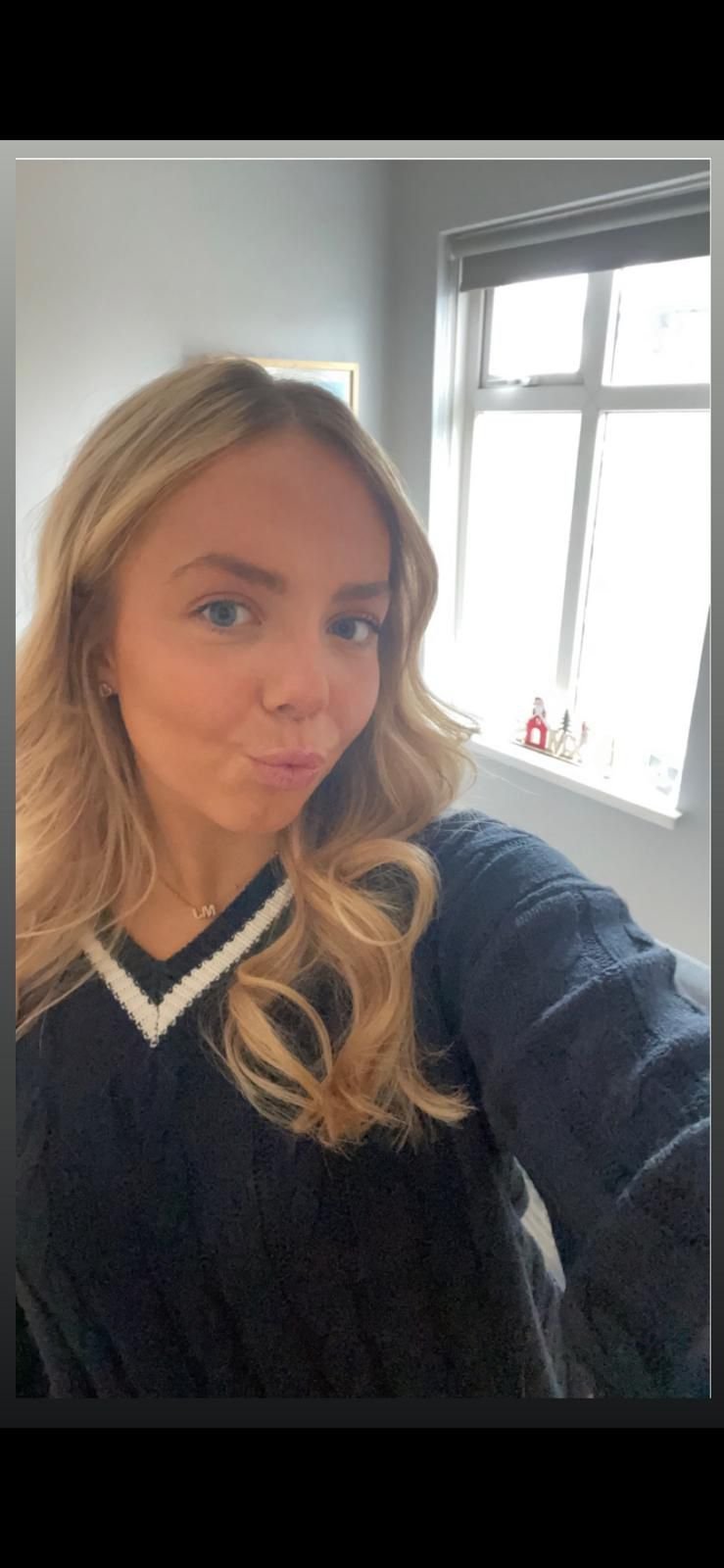 A woman with blonde hair taking a selfie in front of a window in a well-lit room, wearing a dark sweater with a white V-neckline.