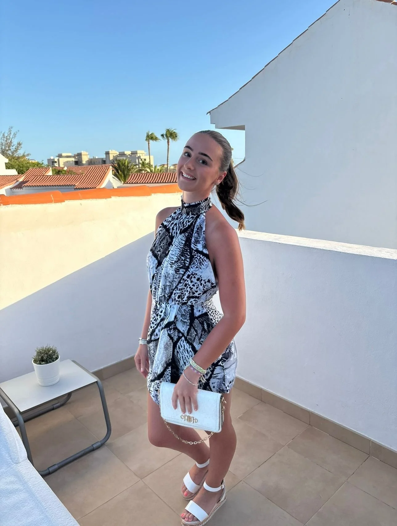 Young woman in a black and white patterned dress holding a white clutch and wearing white sandals on a patio with a white wall and a small table with a potted plant, under a clear blue sky.
