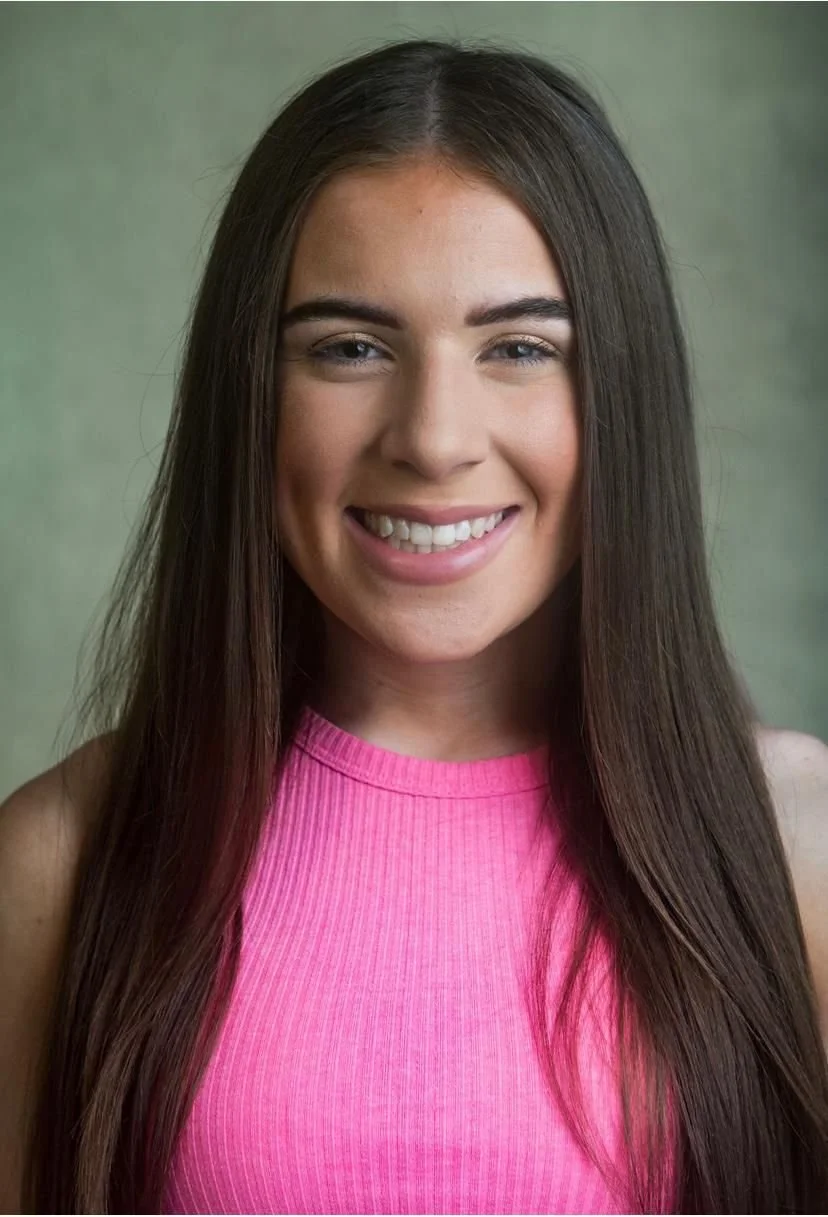 A young woman with long brown hair wearing a pink sleeveless top, smiling with teeth showing, against a plain green background.