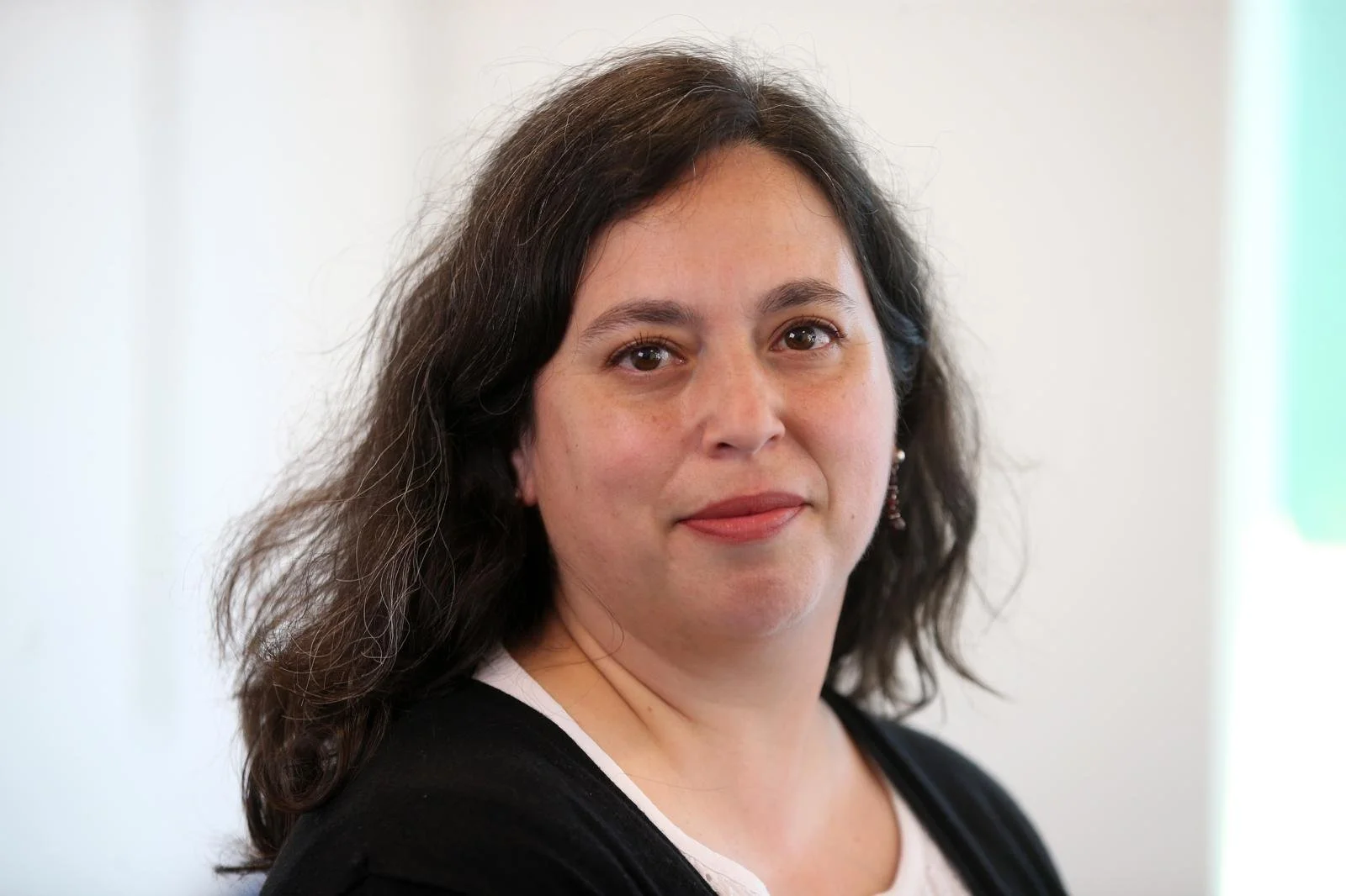 A woman with dark, wavy hair and light skin wearing a white top and black cardigan, looking at the camera with a neutral expression, in an indoor setting with soft lighting.