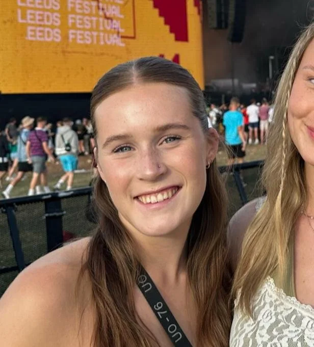 Close-up of a young woman smiling at an outdoor festival, with a crowd and a yellow and red festival sign in the background.