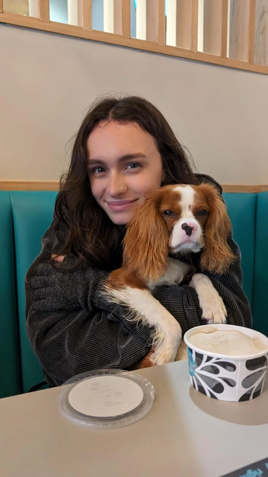 A young woman with dark wavy hair smiling while holding a Cavalier King Charles Spaniel puppy with brown and white fur, sitting at a table in a restaurant with teal booths and wooden accents.