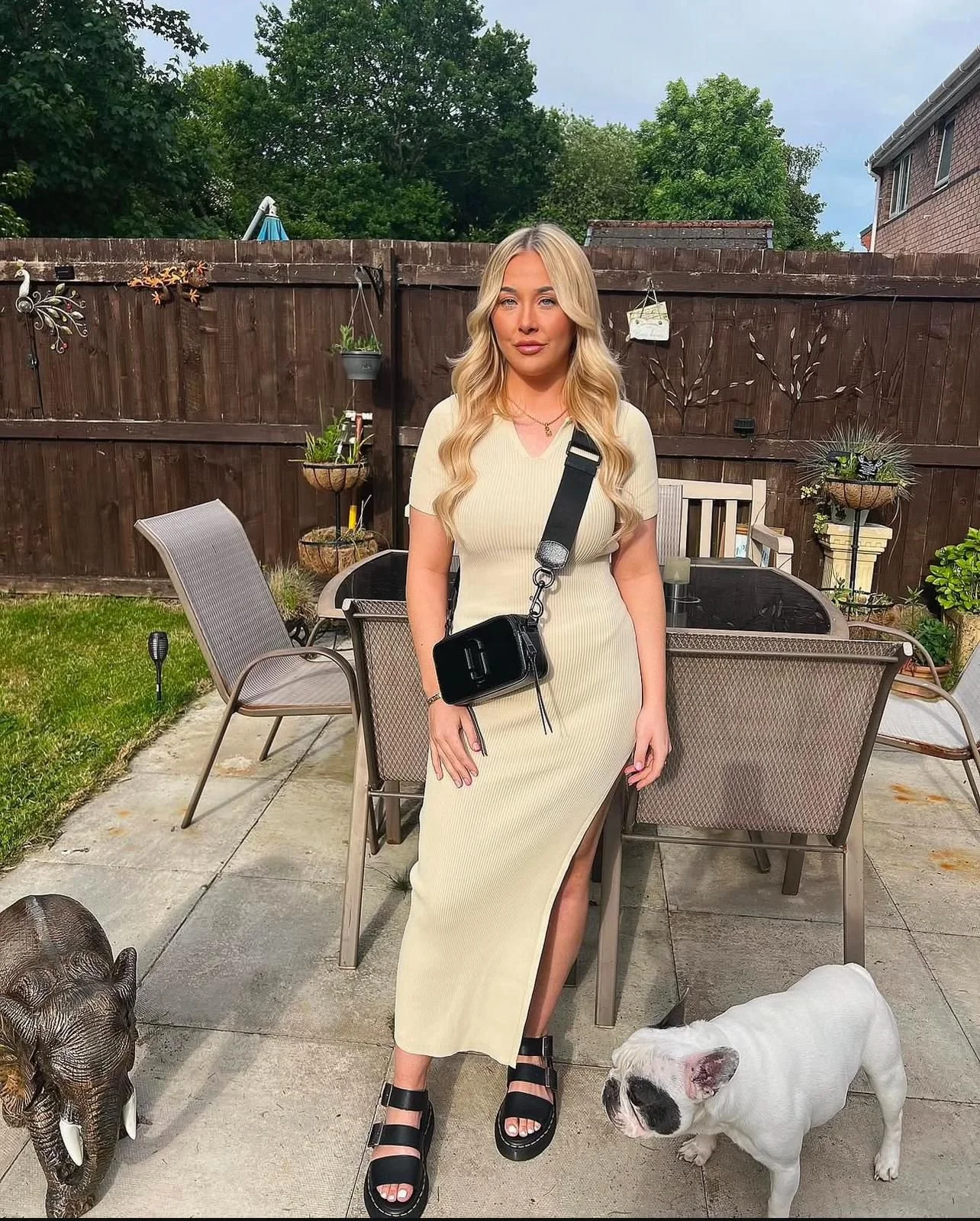 A young woman with long blond hair standing on a patio in a beige dress with a thigh slit. She carries a small black purse with a strap across her body. A white bulldog with black markings sits on the ground to her right. There are outdoor chairs, potted plants, and a wooden fence in the background.
