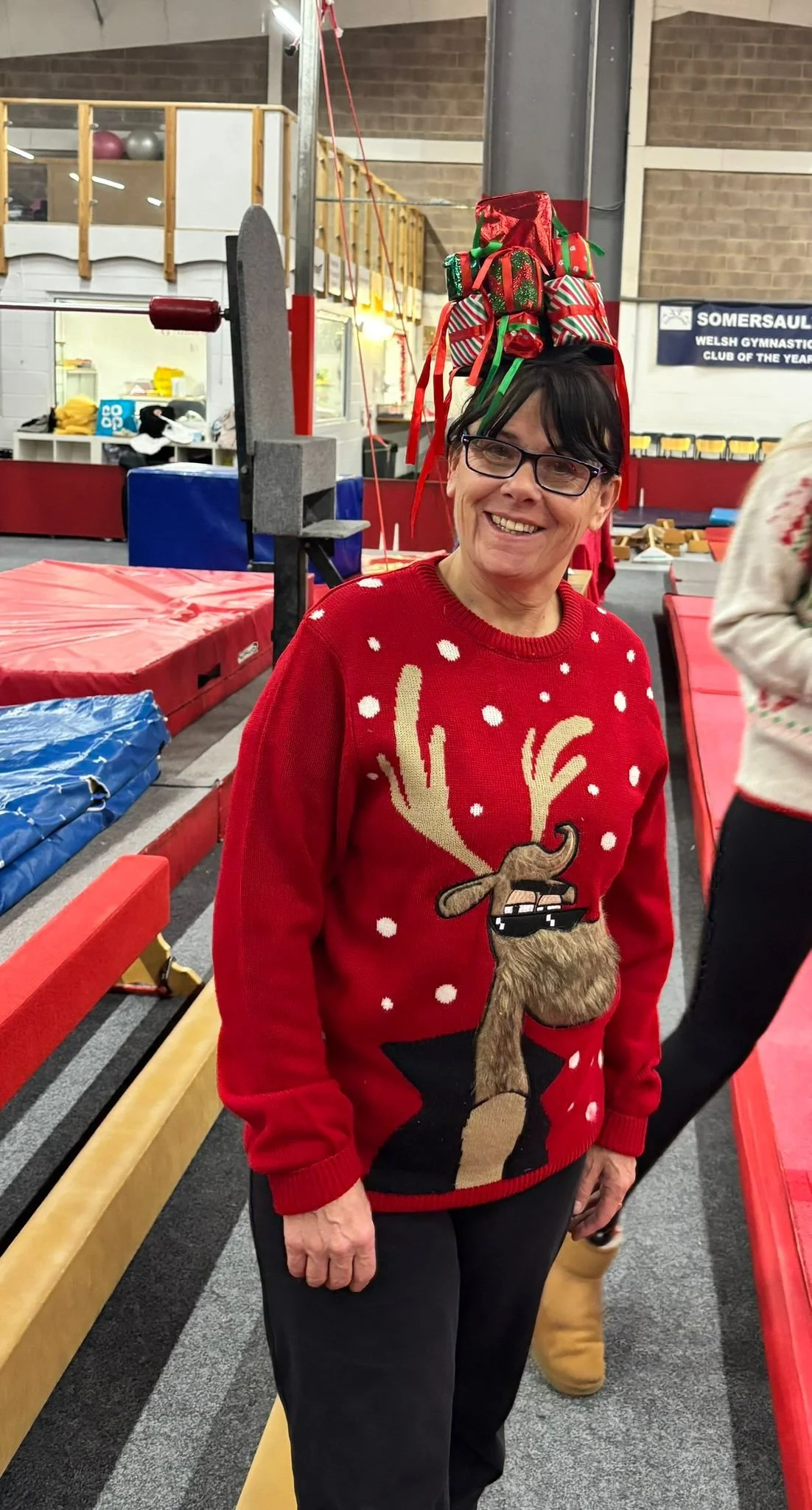 A woman wearing glasses and a red Christmas sweater with a reindeer design standing in a gymnasium, balancing a stack of Christmas stockings on her head.