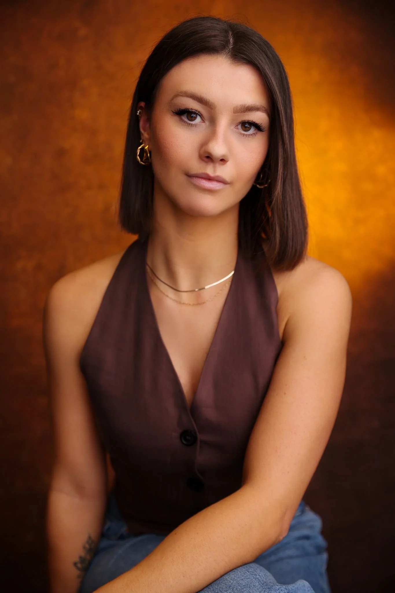 A young woman with shoulder-length dark brown hair, wearing a dark brown sleeveless button-up top, gold hoop earrings, and layered necklaces, sitting against a warm, textured background.