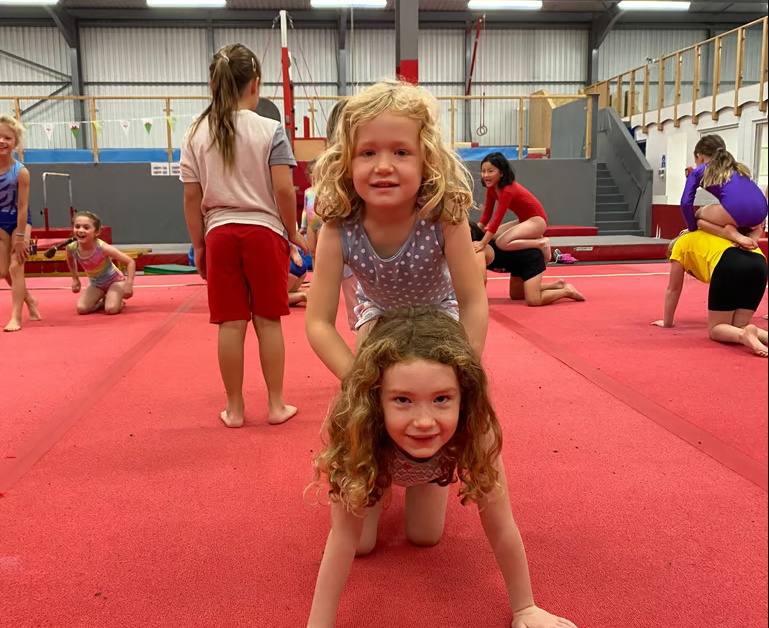 Children practicing gymnastics on a red mat in a gymnasium, with some stretching and others in various poses.