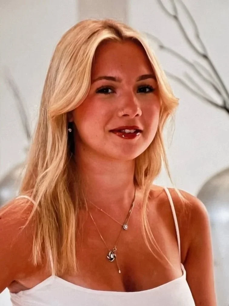 A woman with long blonde hair wearing a white spaghetti strap top, jewelry, and smiling softly.