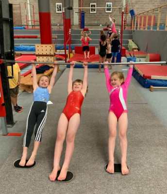 Three young girls hanging from a horizontal bar in a gymnastics gym, with other children and gym equipment in the background.