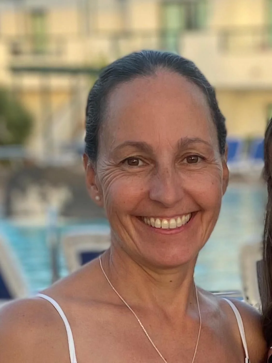 Close-up of a smiling woman with dark hair tied back, wearing a white tank top and a delicate necklace, with a pool and buildings in the background.