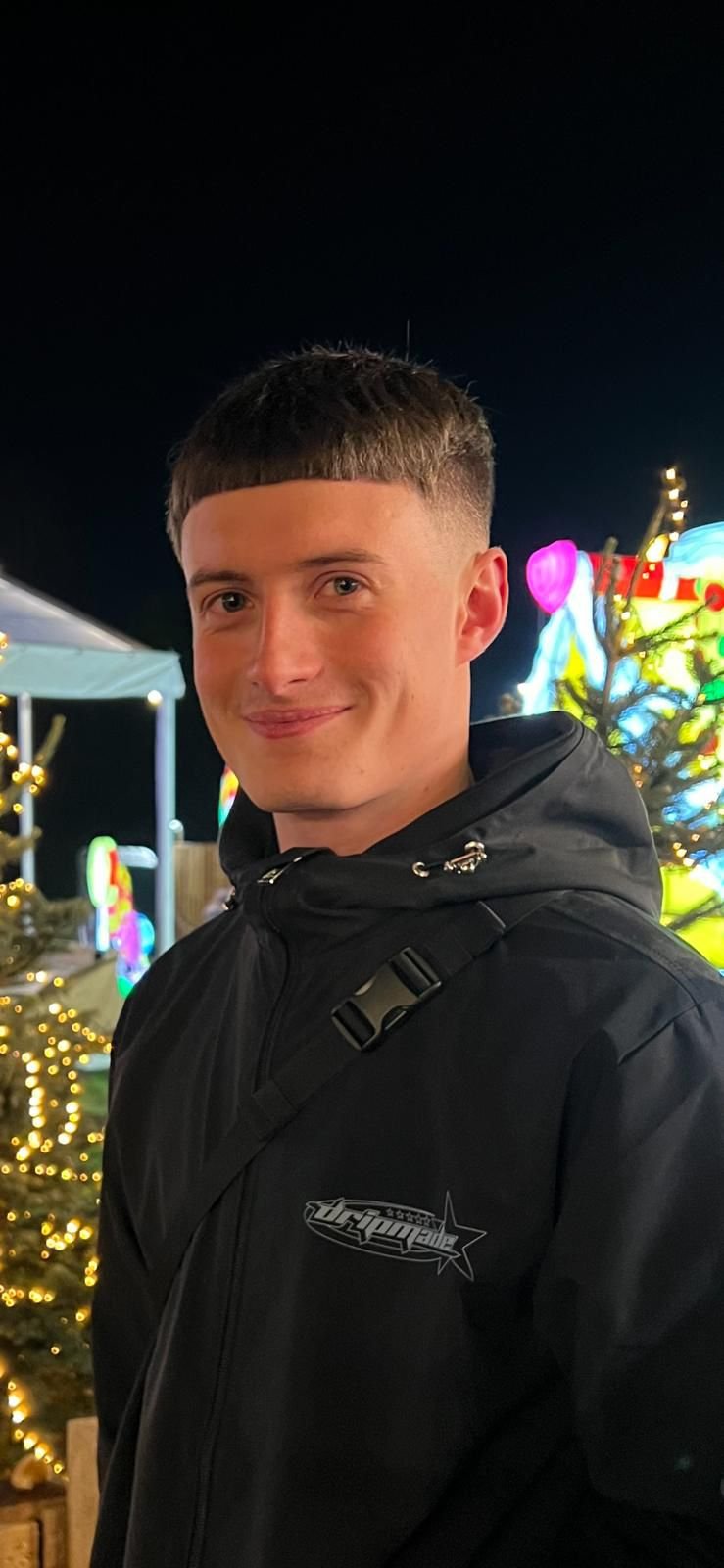 A young man with short brown hair smiling at night, dressed in a black jacket with a backpack, standing in front of holiday lights and decorations.