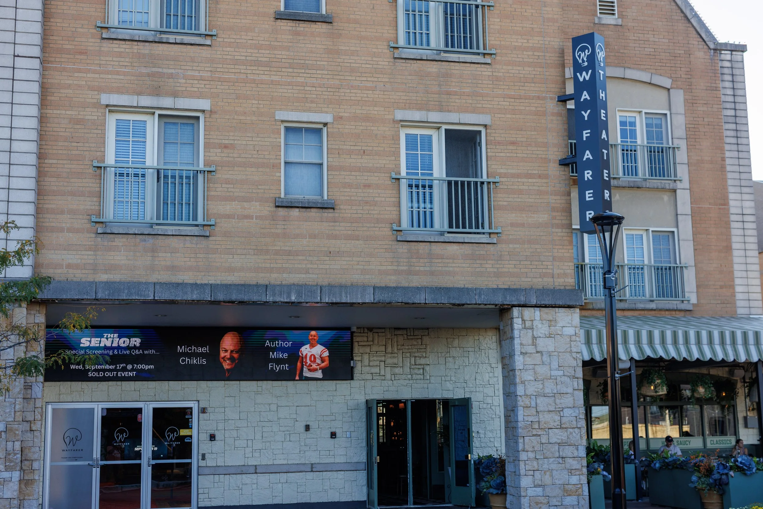 The exterior of Wayfarer Theatre, a multi-story building with brick and stone facade, featuring a vertical blue sign with the theater's name and logo, and an electronic marquee promoting an event with Michael Chiklis and Mike Flynt.