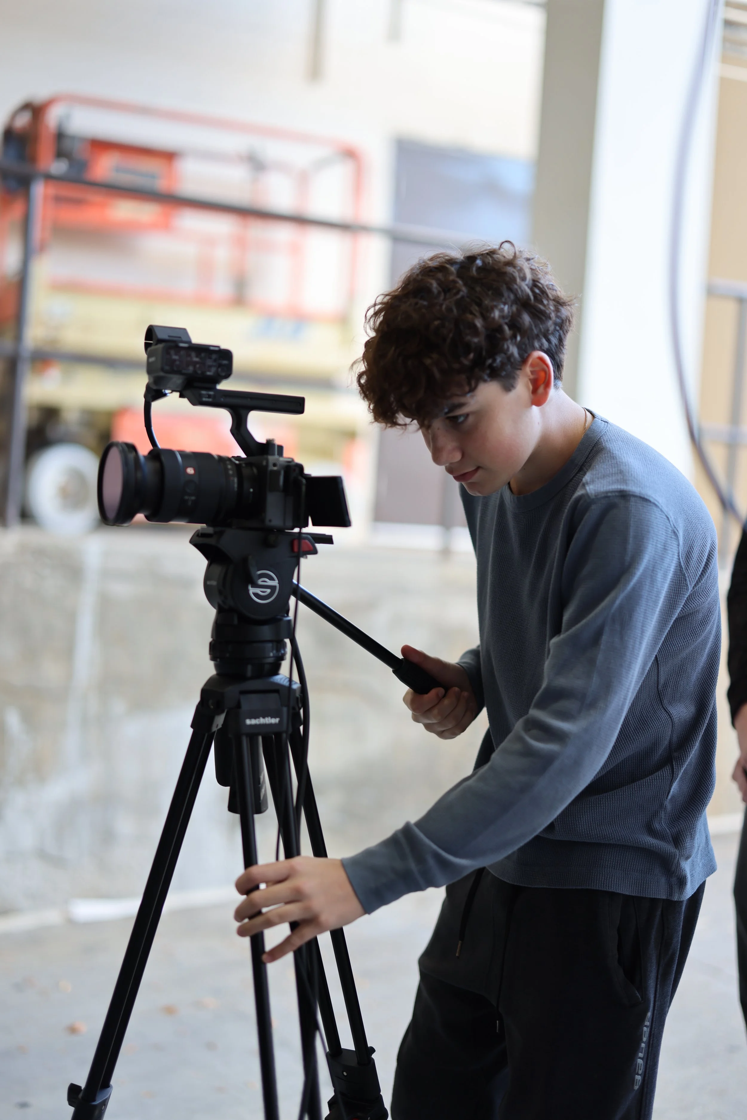 A young boy with curly hair adjusting a professional video camera on a tripod indoors.