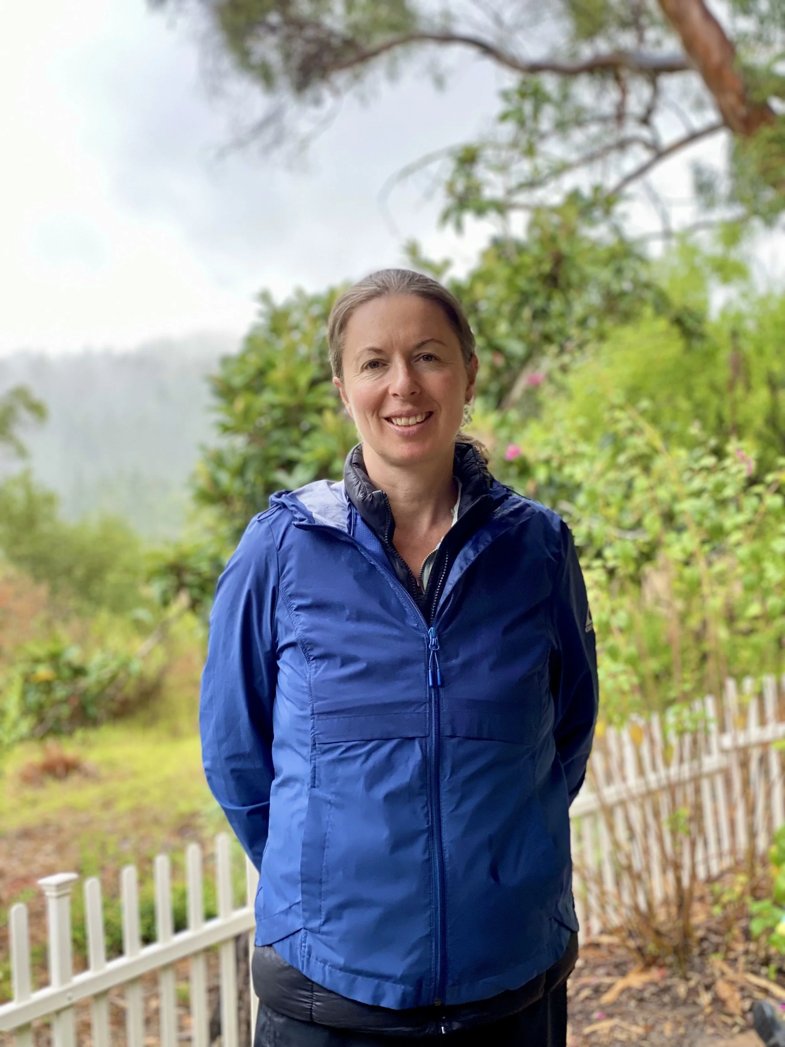 Woman in a blue jacket smiling outdoors with trees and a white picket fence in the background.