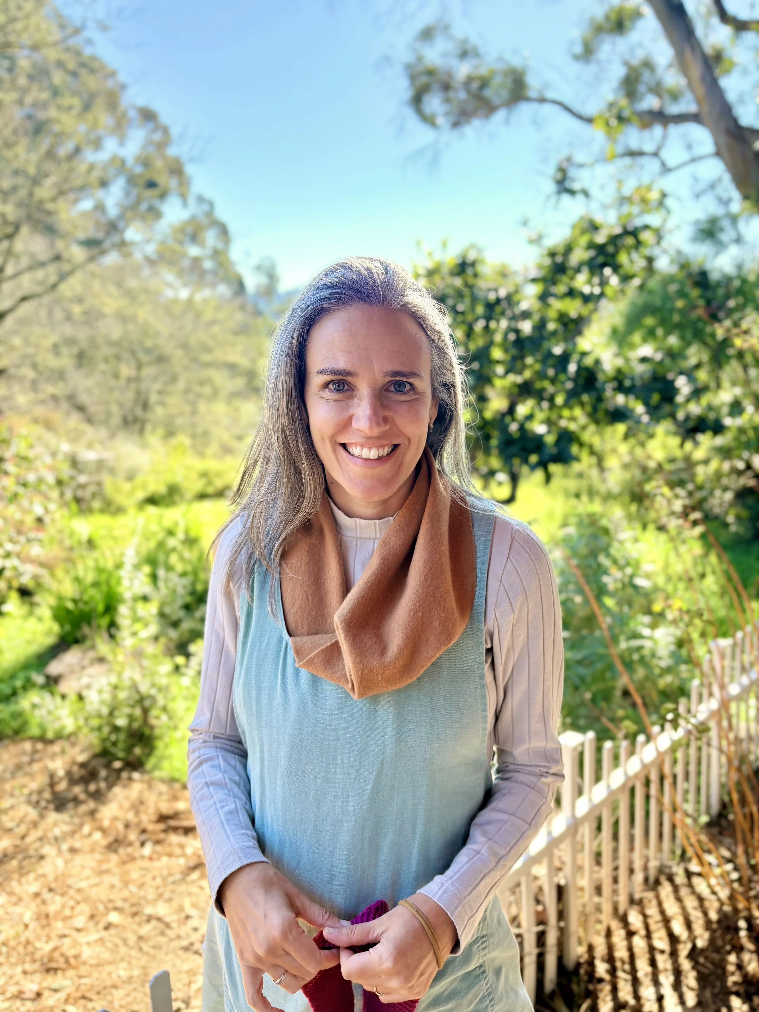 A woman outdoors smiling, with trees and greenery in the background on a sunny day.
