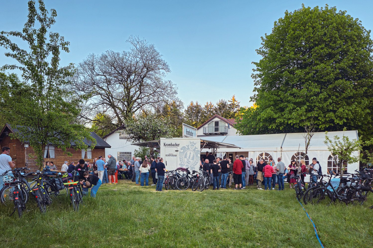 Menschen versammeln sich vor einer weißen Zeltabbund auf einer grünen Wiese. Fahrräder sind am Rand geparkt, viele Bäume im Hintergrund, einige Häuser. Es sieht nach einer Veranstaltung oder einem Fest aus.