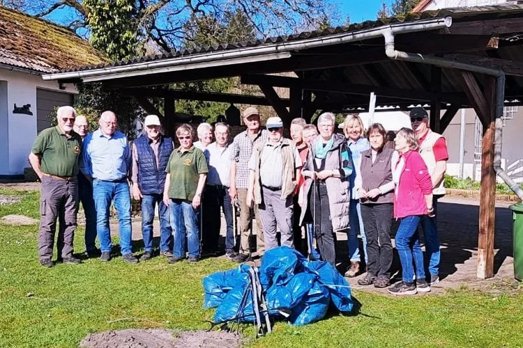Gruppe von Menschen steht im Freien vor einem Holzpavillon, in der Nähe liegen blaue Müllsäcke, vermutlich bei einer Reinigungsaktion.