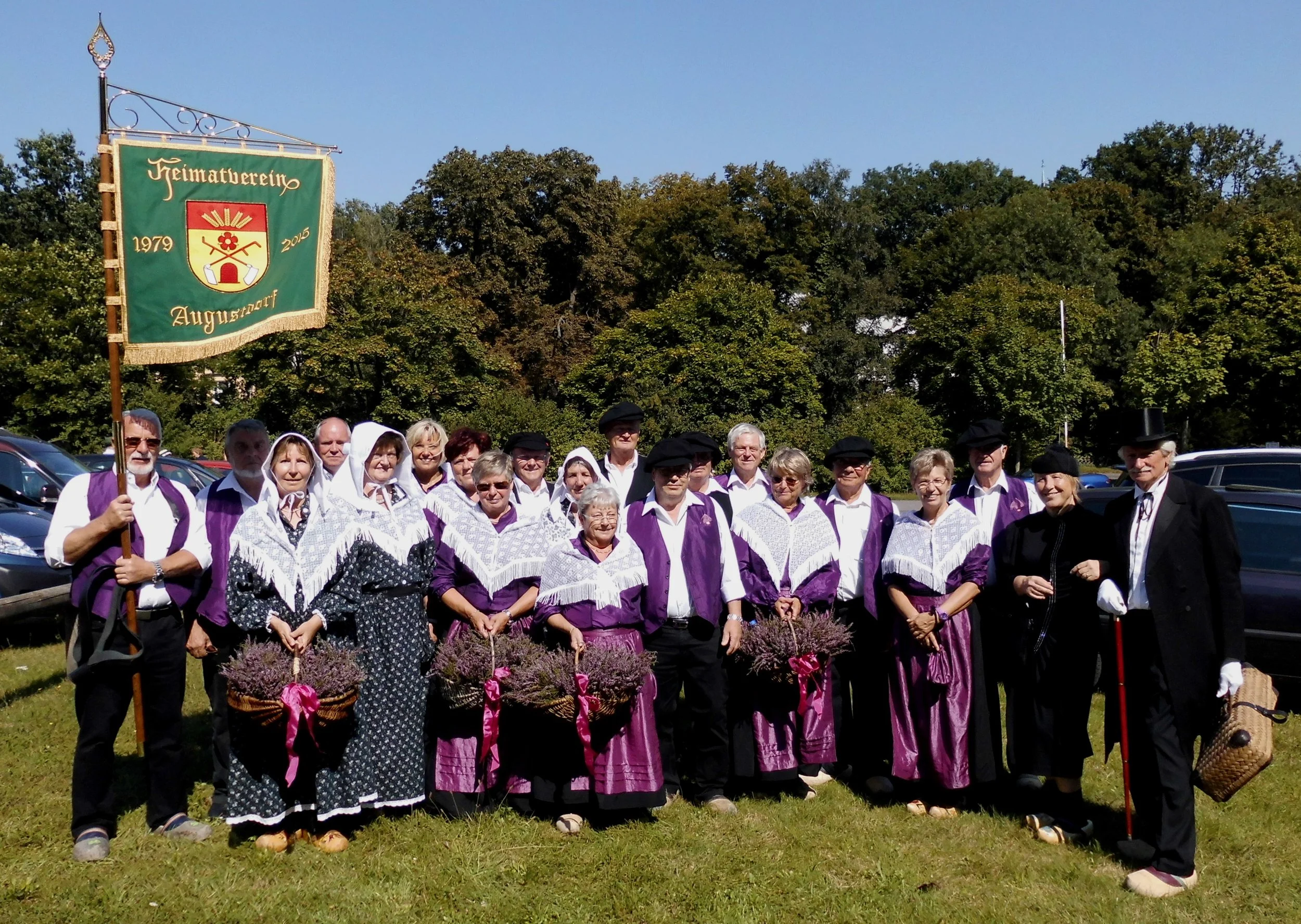 Gruppen von Menschen in traditioneller Kleidung vor einem Baum mit einem grünen Fahnenmast, auf dem steht 'Heimatverein Augustdorf 1979-2015'.