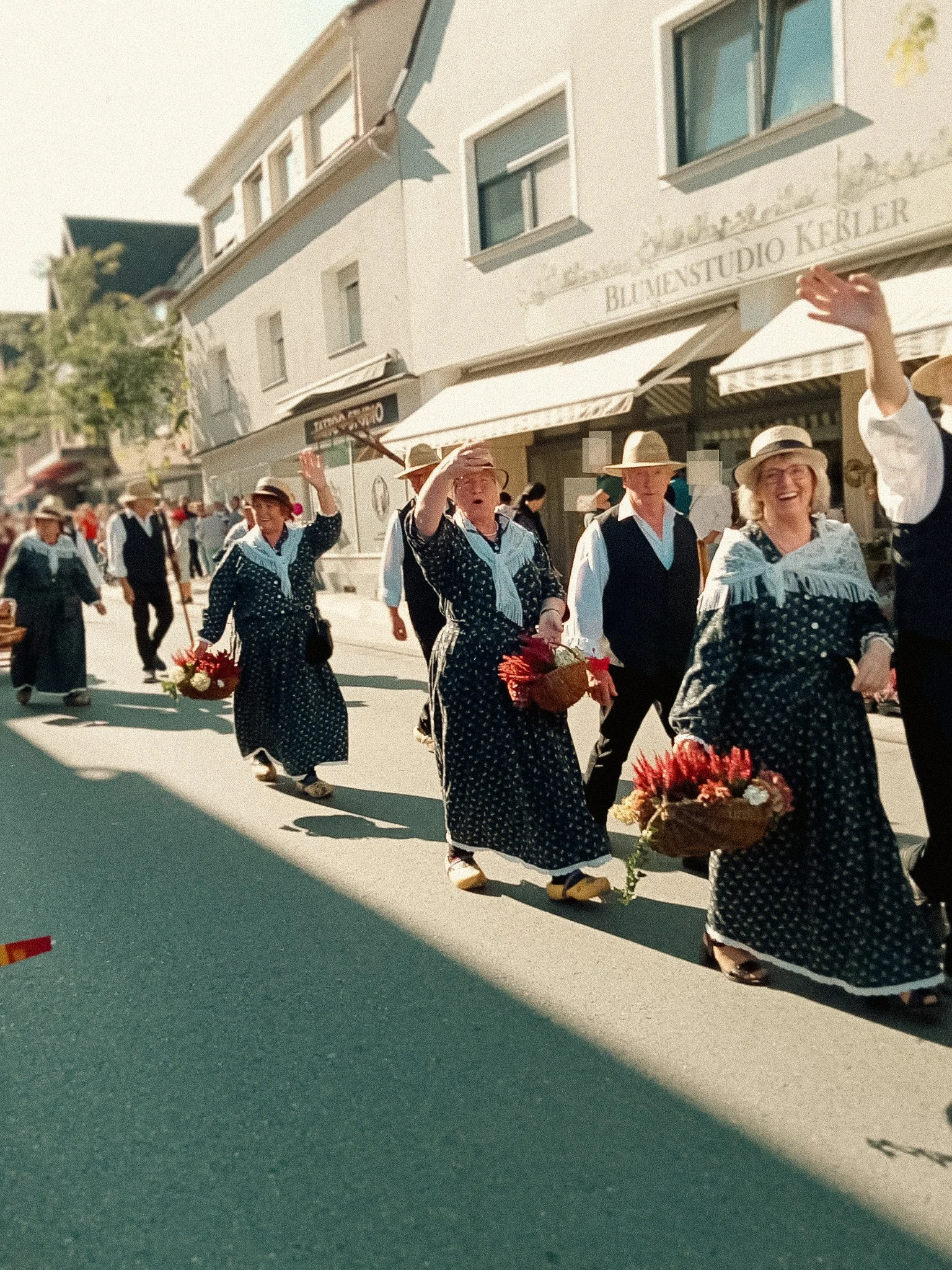 Menschen in traditioneller Kleidung beim Umzug, einige tragen Hüte, einige Frauen haben Blumensträuße, auf einer Straße in einer kleinen Stadt oder einem Dorf.