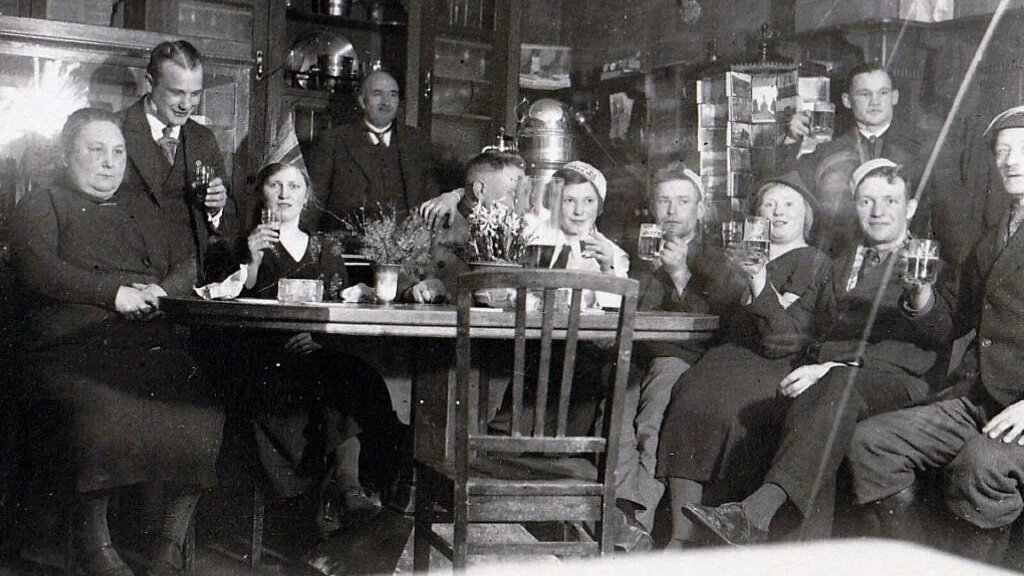 Eine Gruppe von Menschen in einem rustikalen Raum, alle sitzen oder stehen um einen Tisch, einige halten Biergläser hoch, vermutlich bei einem geselligen Beisammensein in der Vergangenheit.