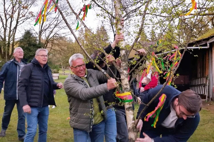 Eine Gruppe von Menschen bei einer Baumzeremonie im Freien, die den Baum mit bunten Bändern und Quasten schmücken.