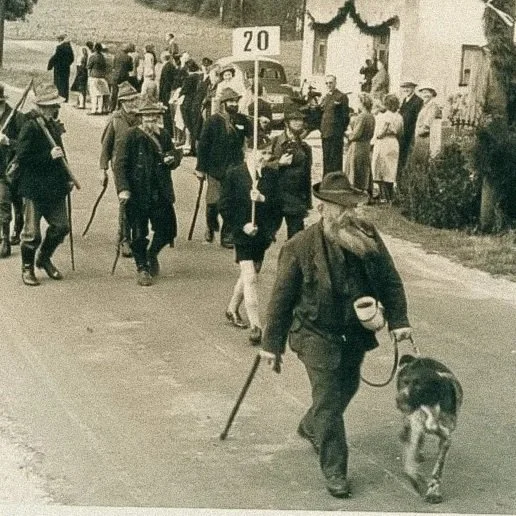 Menschen auf einer Straße, einige mit Hüten, ein Mann mit Hund und Stock läuft im Vordergrund; im Hintergrund Menschen vor Gebäuden.