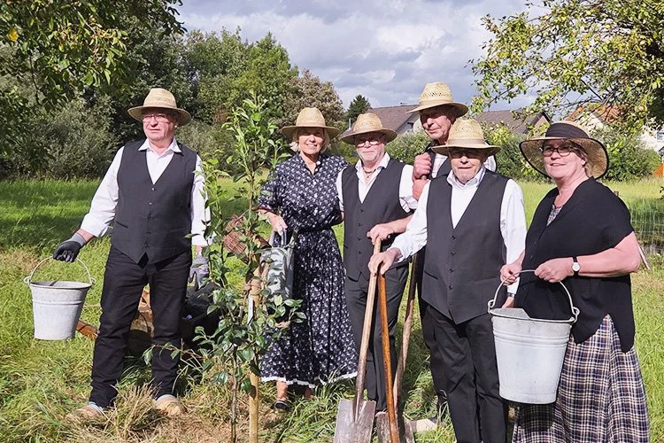 Sechs Menschen, größtenteils älter, stehen im Garten mit Gummihandschuhen, Hüten und Gartengeräten, um einen jungen Baum zu pflanzen