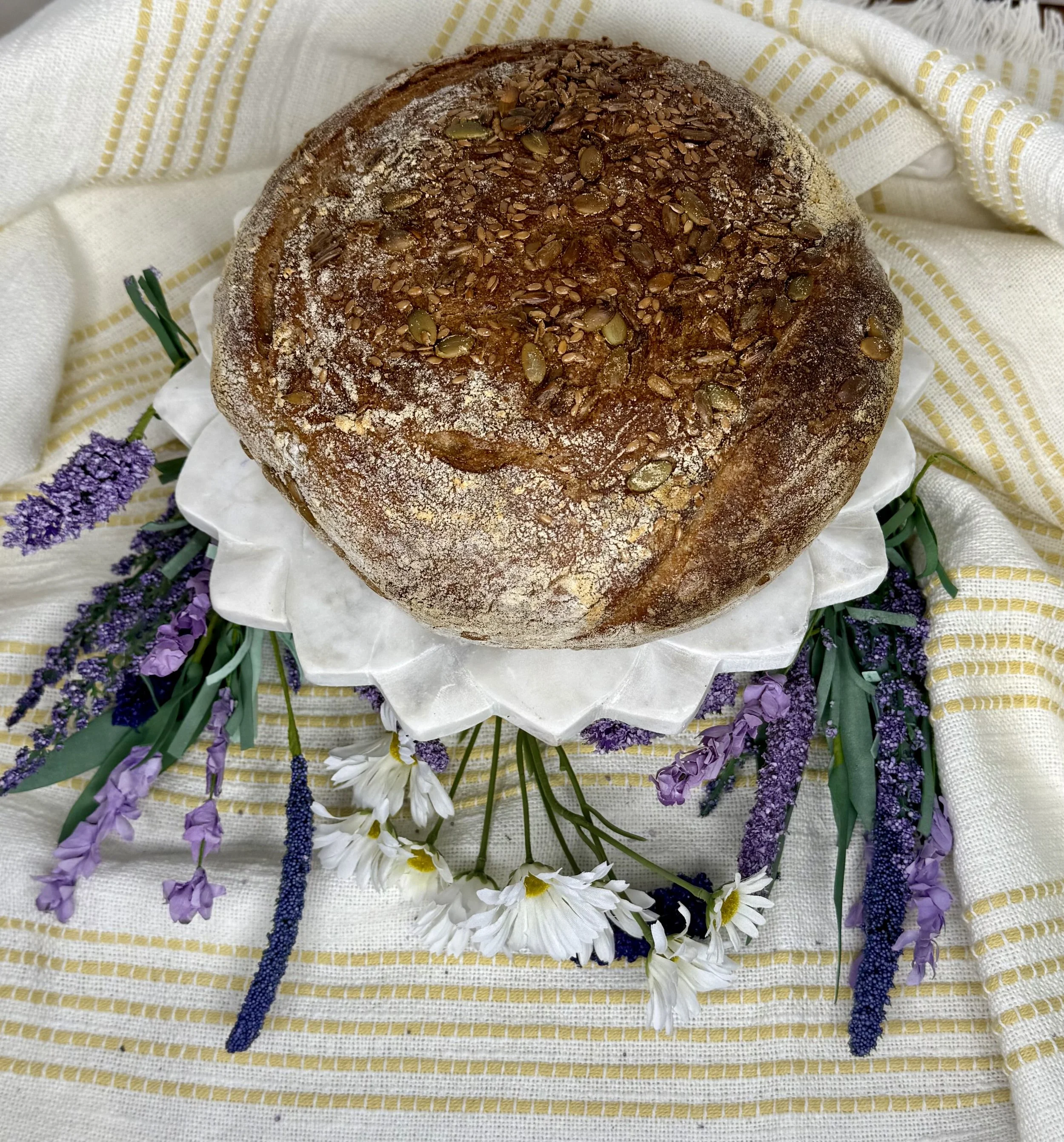 A round loaf of bread topped with pumpkin seeds, placed on a white plate with ruffled edges, surrounded by purple and white flowers on a striped yellow and white cloth.