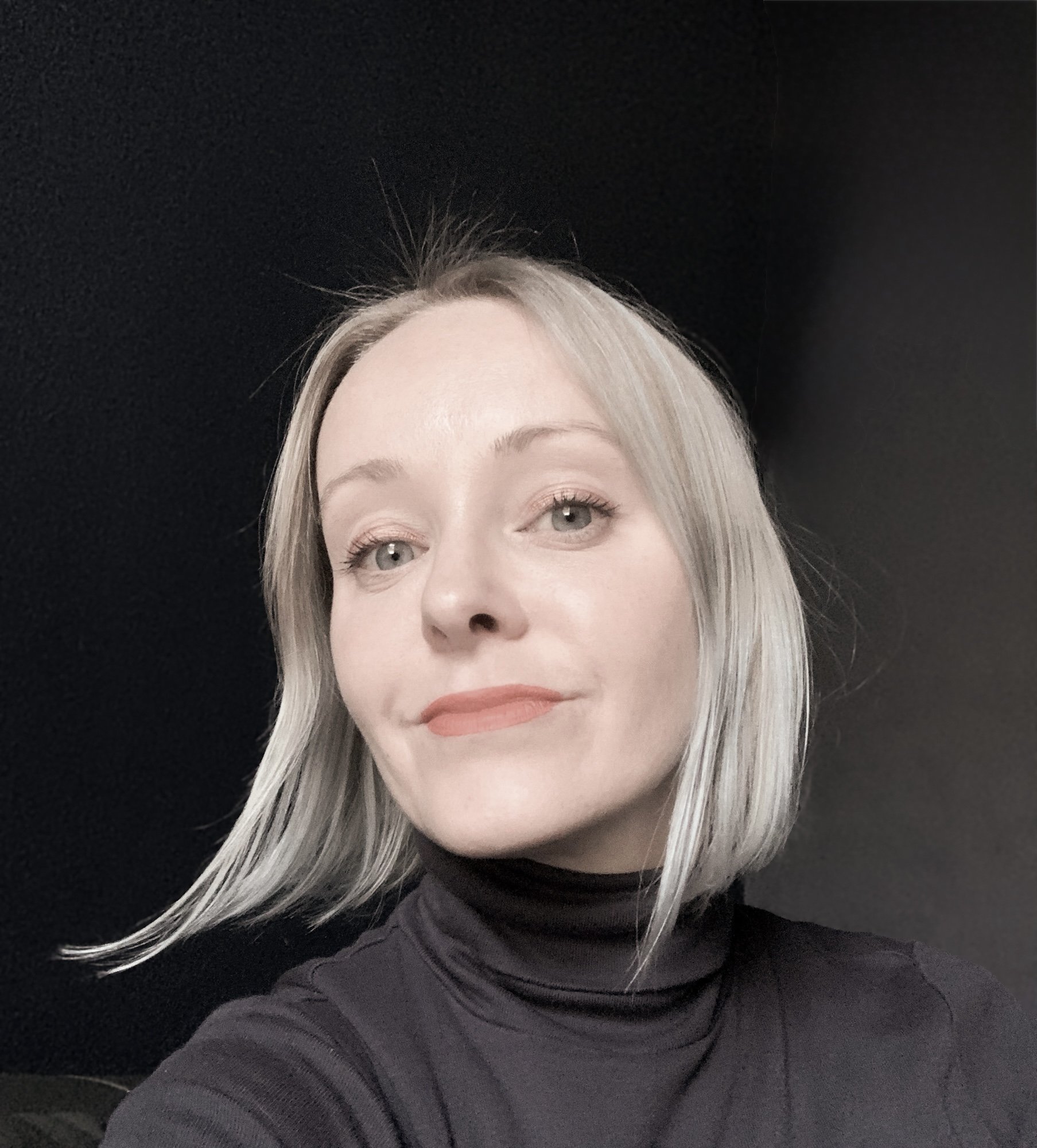 A woman with blonde hair and light skin wearing a black turtleneck posing for a profile photograph against a dark background.