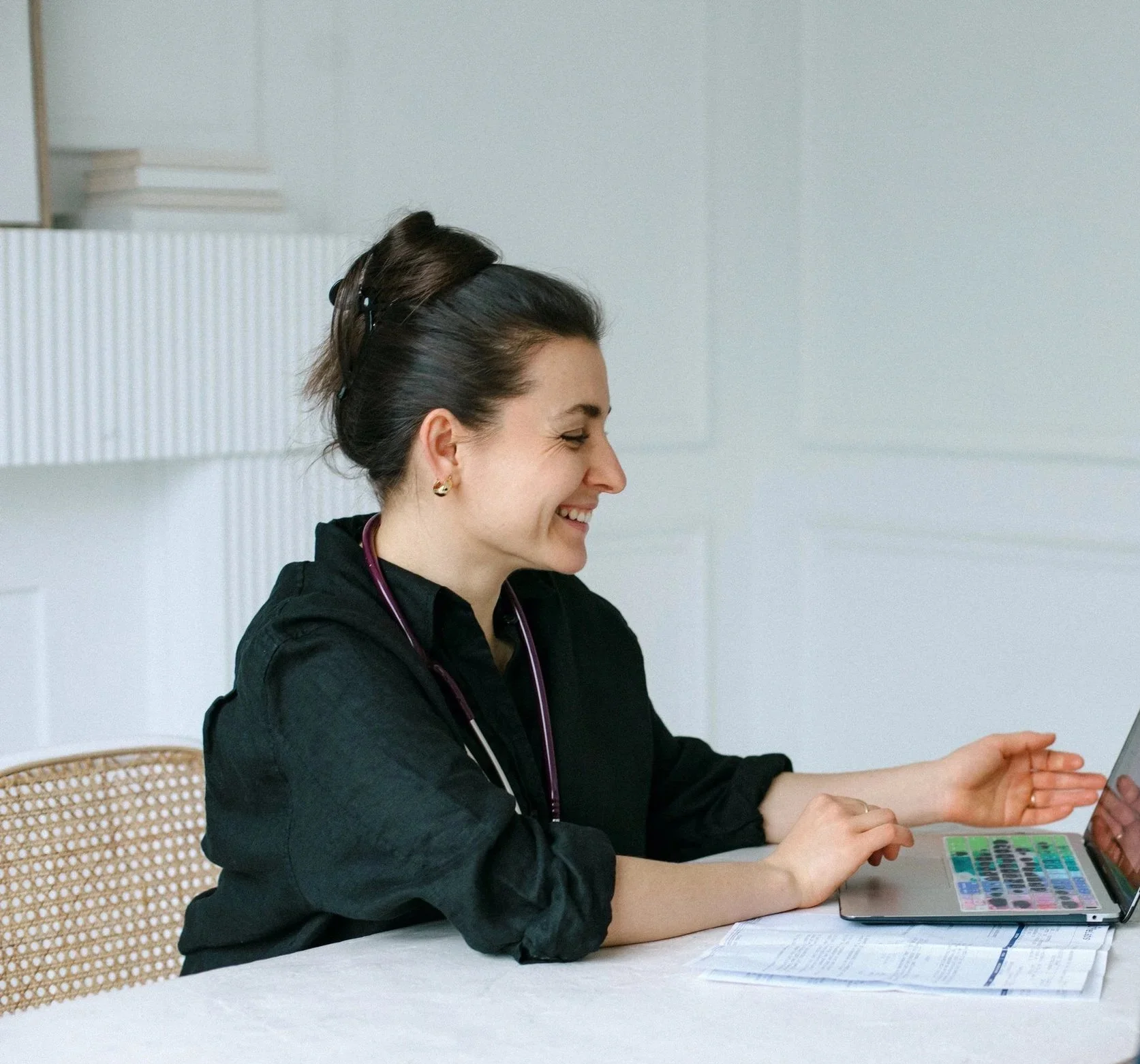 A woman with dark hair in a bun, smiling and looking at a tablet. She is wearing a black shirt, earrings, and a stethoscope around her neck, sitting at a white table with papers and a colorful chart or calendar.