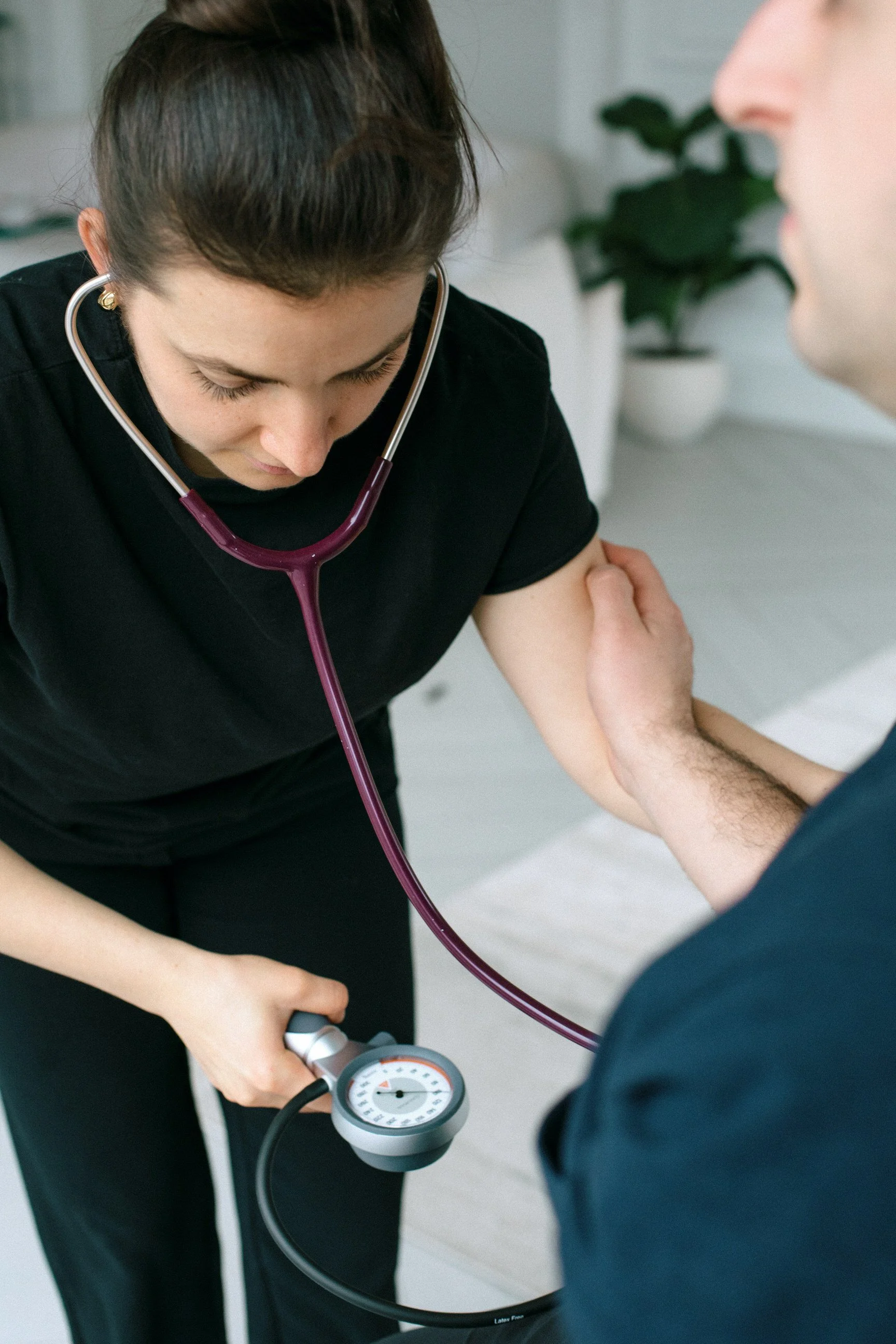 A healthcare professional, likely a nurse or doctor, is measuring a patient's blood pressure with a sphygmomanometer. The professional is wearing a black uniform and has a stethoscope around her neck, focusing on the blood pressure cuff on the patient's arm. The scene is in a clinical setting with a plant in the background.