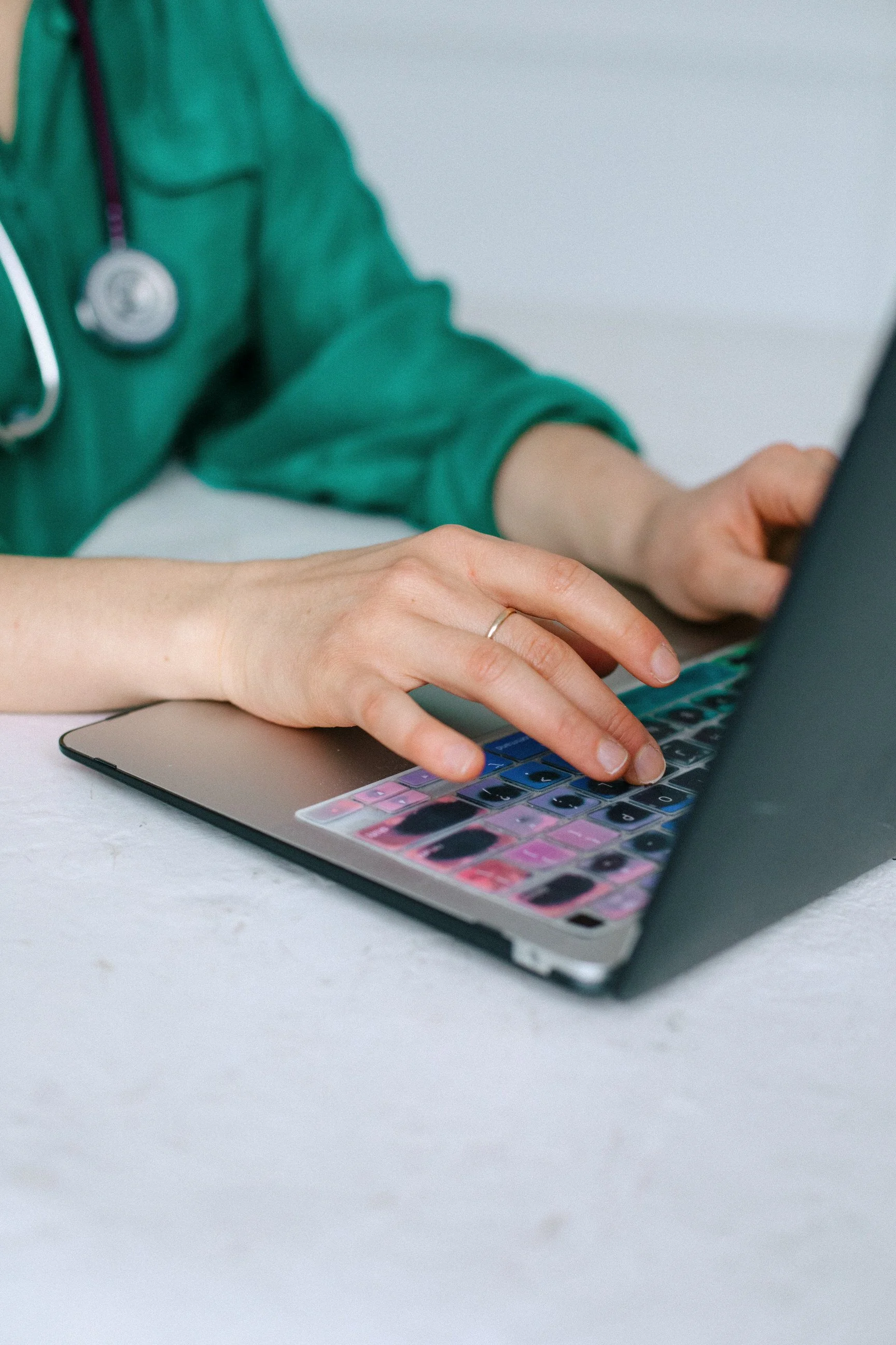 A person in green medical scrubs with a stethoscope around their neck is typing on a colorful laptop keyboard on a white table.
