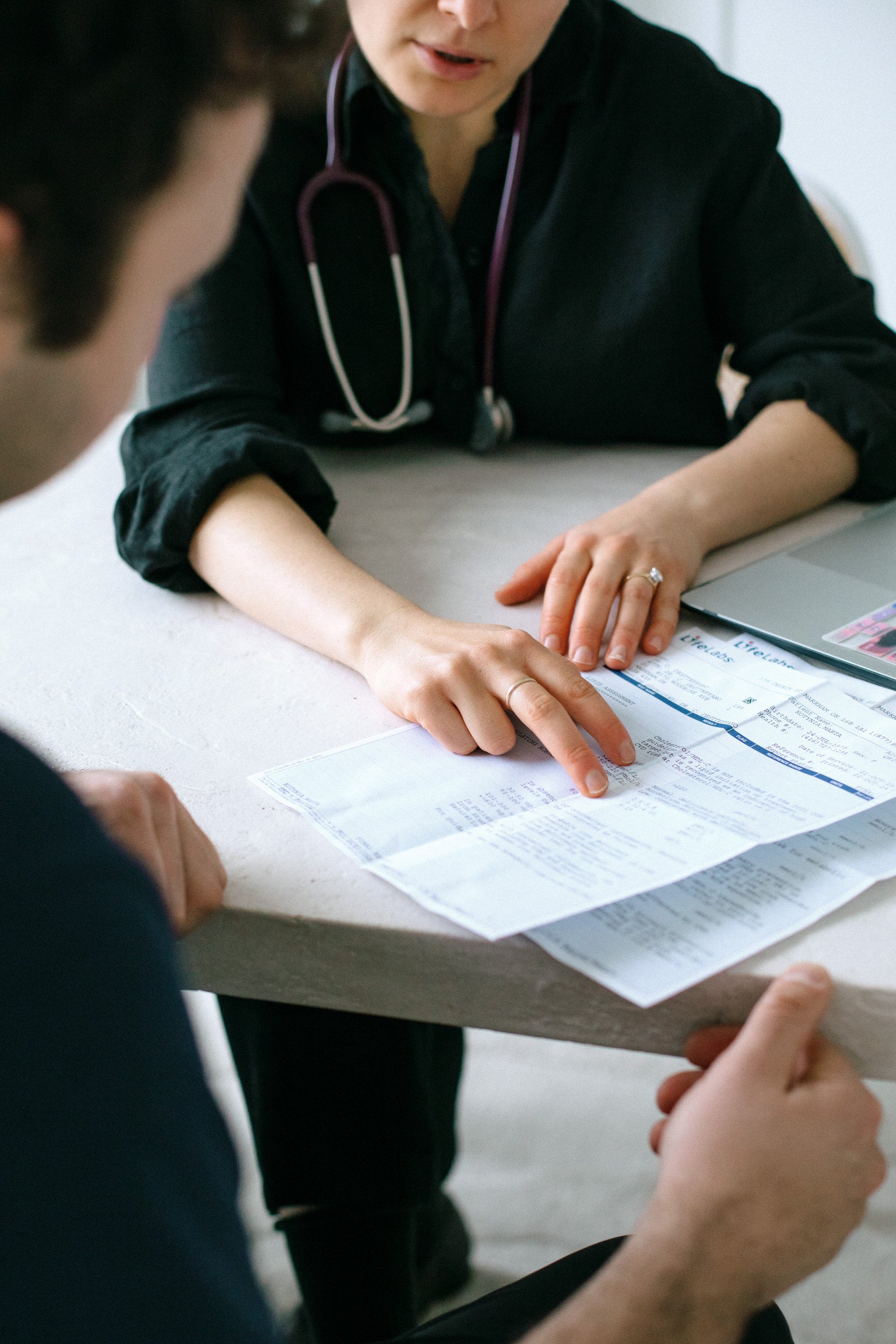 A healthcare professional with a stethoscope around her neck is pointing to a document on a table, explaining information to a patient. The patient is listening attentively, with hands visible on the table.