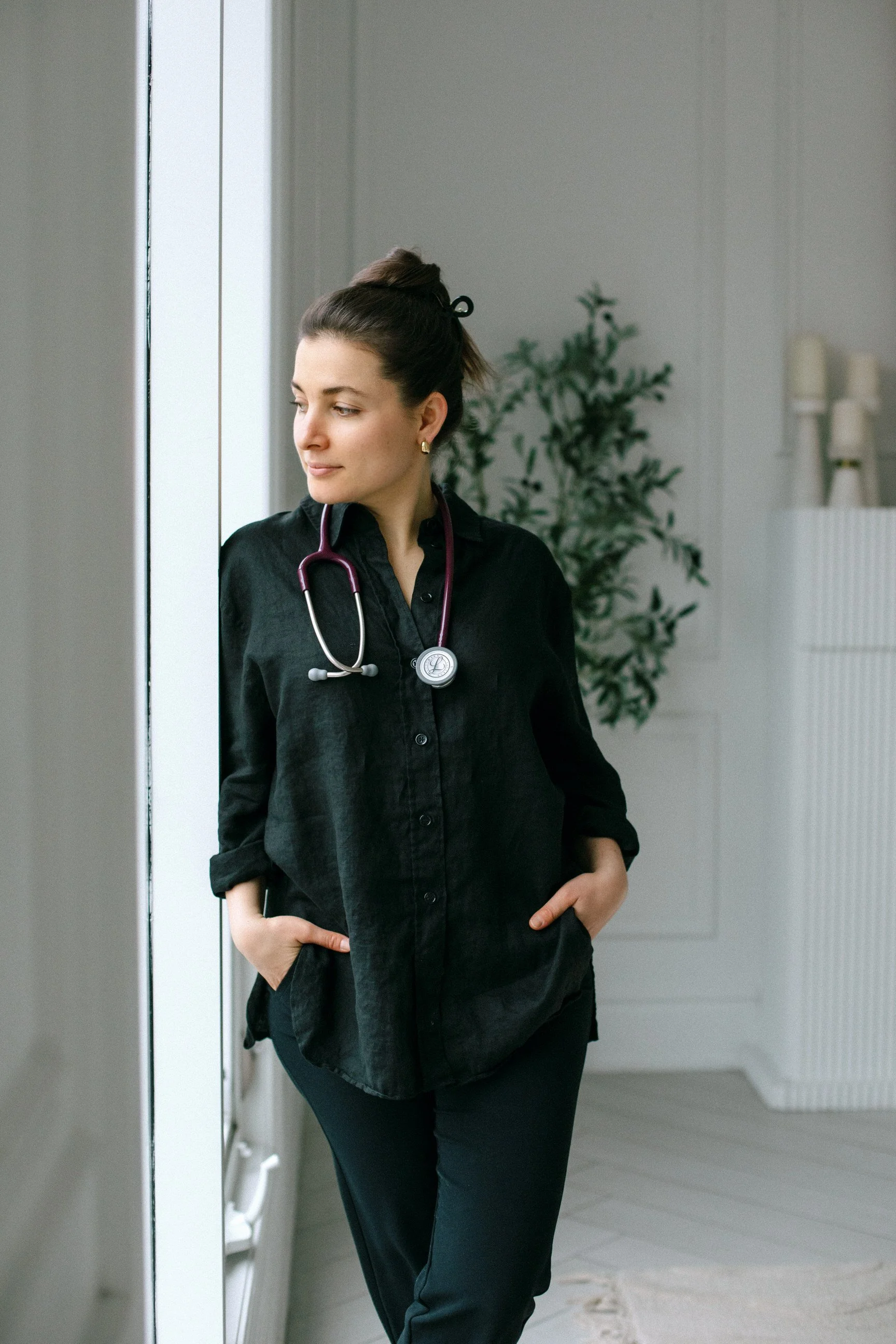 A woman with dark hair in a bun wearing a black shirt with a stethoscope around her neck, standing in a room with light-colored walls and a plant in the background.