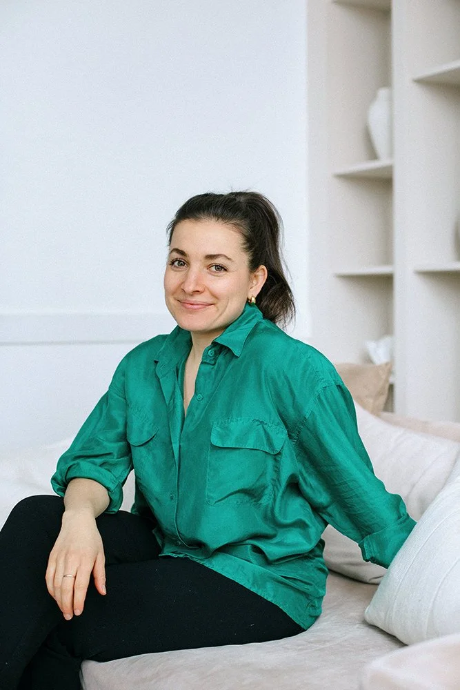 A woman sitting on a bed, smiling, wearing a teal button-up shirt and black pants, in a bedroom with white walls and built-in white shelves.