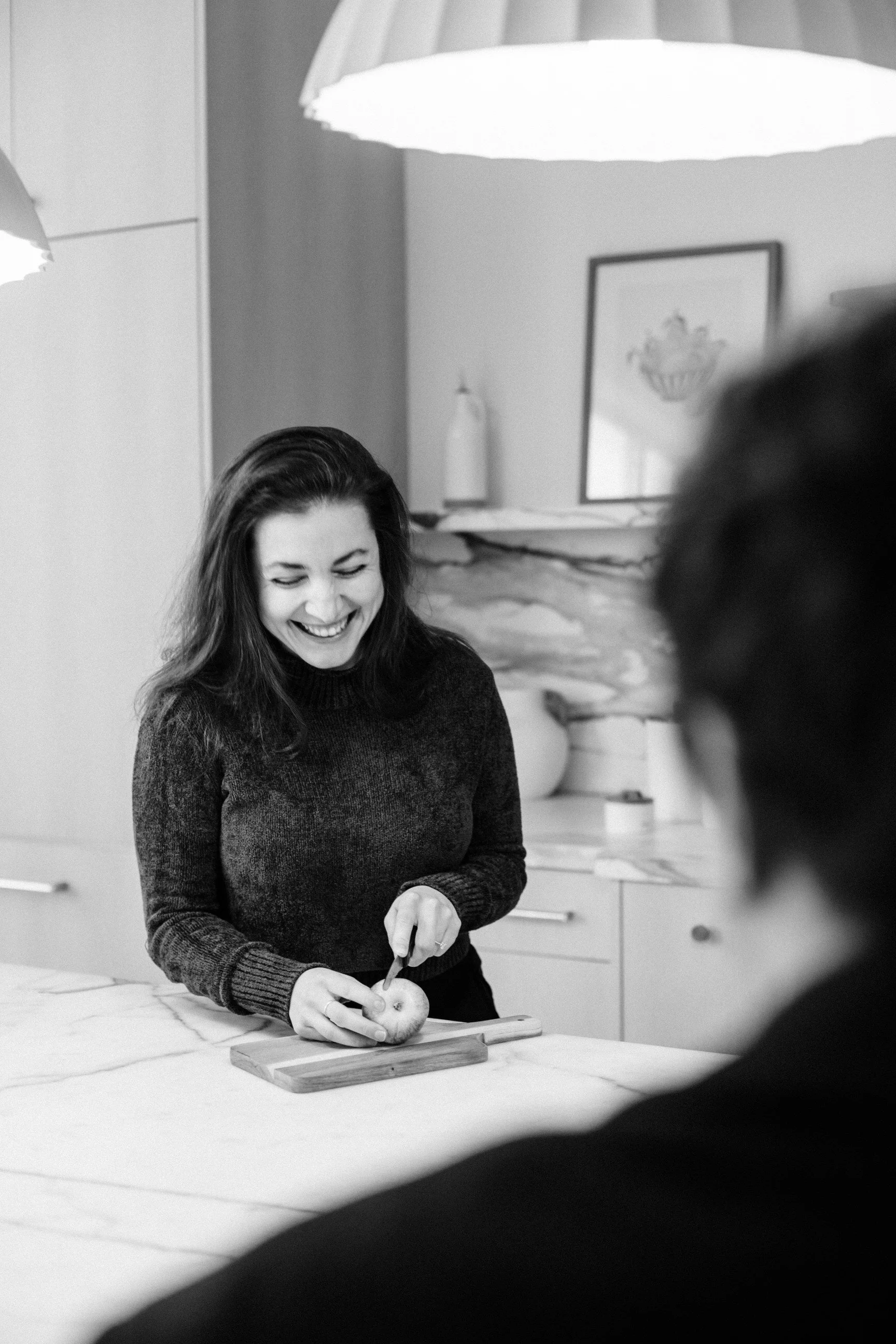 A woman with long dark hair smiling while slicing an apple on a cutting board in a kitchen, with a framed picture and a lamp in the background.
