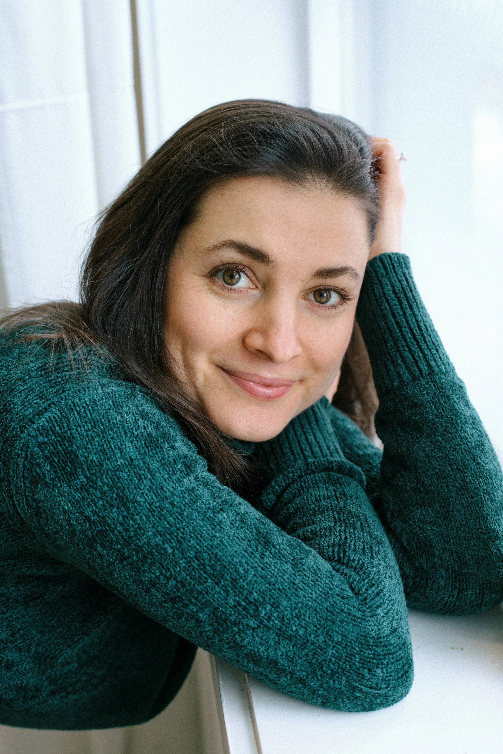 A young woman with dark brown hair and green eyes, wearing a green sweater, smiling gently, resting her head on her hand, sitting at a white table near a window with white curtains.